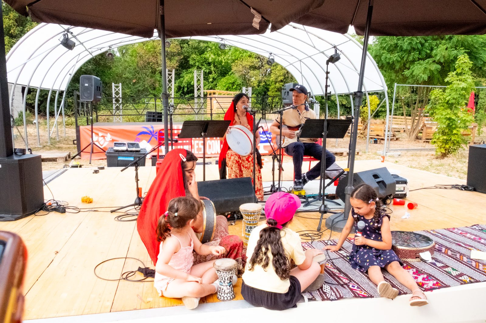 Scène sous chapiteau, enfants assis sur tapis face aux musiciens